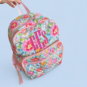 May include: A quilted backpack featuring a vibrant floral pattern with pink, yellow, and green flowers on a light blue background. The bag includes a front zippered pocket and a large main compartment, both with pink trim and hot pink monogrammed letters EES.