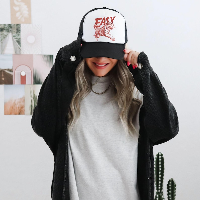 Puede incluir: Gorra de camionero en blanco y negro con la palabra "EASY" en letras rojas y un gr&aacute;fico de tigre rojo. La gorra tiene una parte trasera de malla y una visera curva.