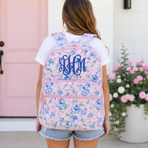 May include: A quilted backpack featuring a light pink background with a blue floral pattern. The bag has a front zippered pocket and a dark blue monogram with three stylized letters centered on the upper portion of the main compartment.