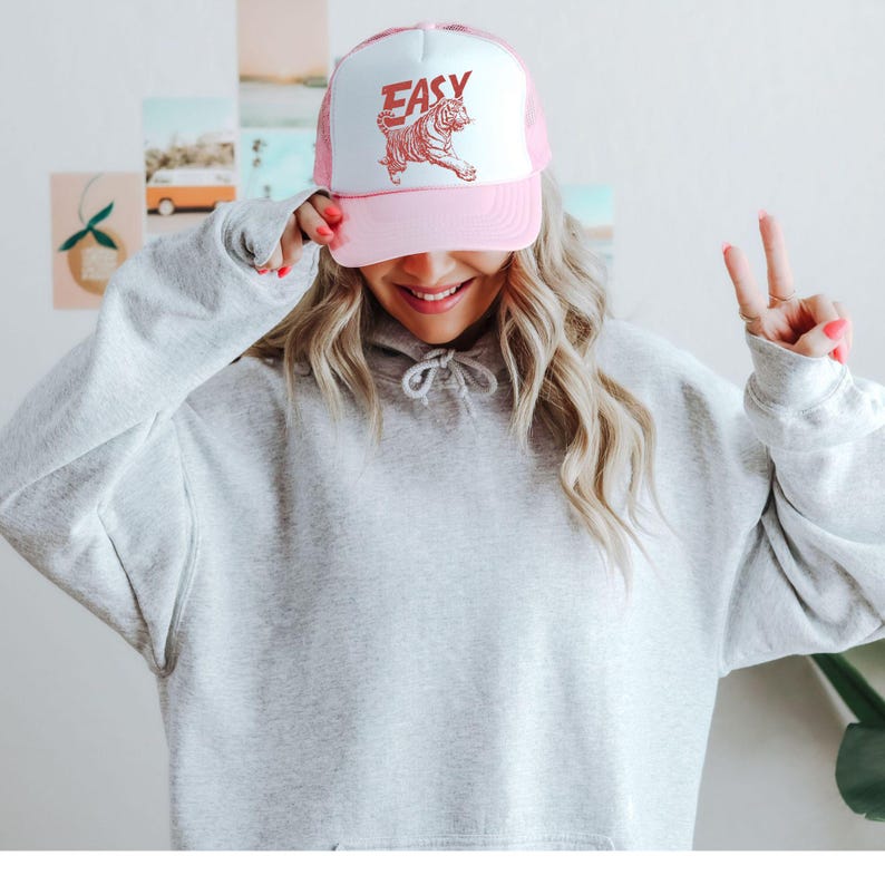 Puede incluir: Gorra de camionero rosa y blanca con la palabra "EASY" en letras rojas y un gr&aacute;fico de tigre rojo. La gorra tiene una parte trasera de malla y una visera curva.