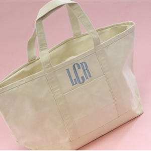 May include: A large, off-white canvas tote bag with a single pocket on the front. The bag has two handles and the initials "LCR" are embroidered in blue on the pocket.