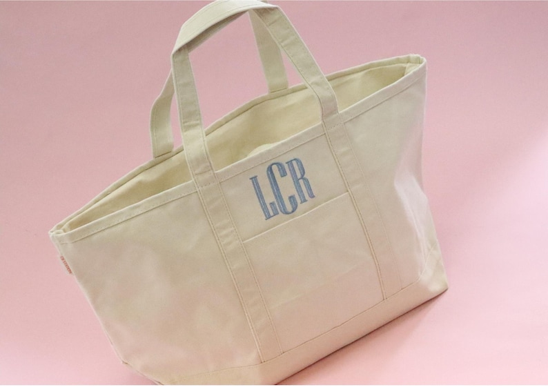 May include: A large, off-white canvas tote bag with a single pocket on the front. The bag has two handles and the initials "LCR" are embroidered in blue on the pocket.