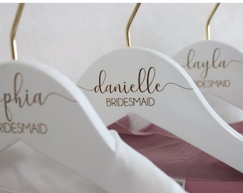 May include: Three white wooden hangers with gold hooks. Each hanger has a different name written in gold script: "Sophia Bridesmaid", "Danielle Bridesmaid", and "Layla Bridesmaid".