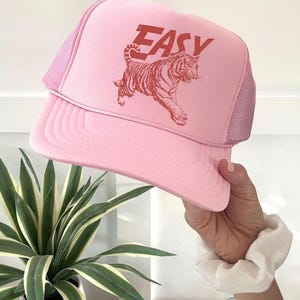 Puede incluir: Gorra trucker rosa con parte trasera de malla y visera curva. La gorra presenta la palabra "EASY" en letras rojas en bloque sobre un gr&aacute;fico de tigre rojo. La gorra es sostenida por una mano.