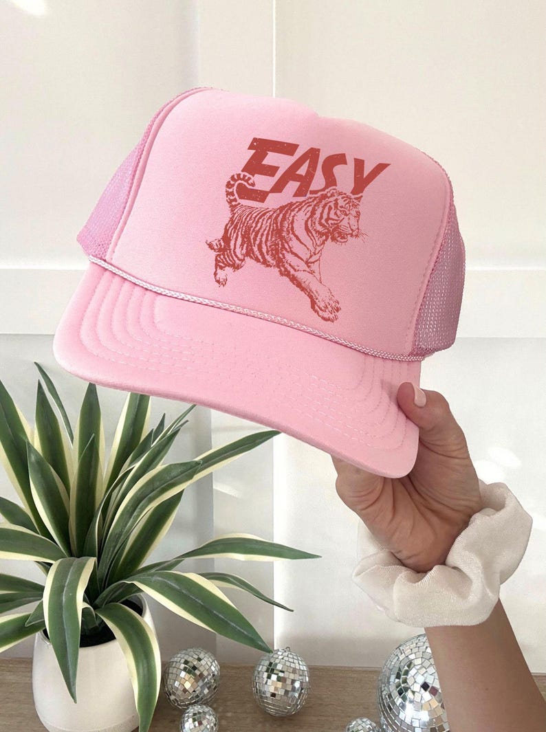 Puede incluir: Gorra trucker rosa con parte trasera de malla y visera curva. La gorra presenta la palabra "EASY" en letras rojas en bloque sobre un gr&aacute;fico de tigre rojo. La gorra es sostenida por una mano.