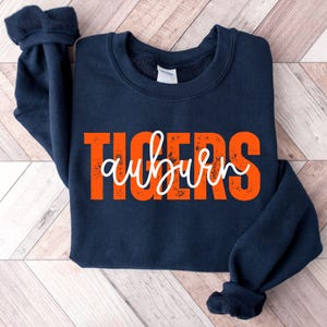 May include: Navy blue sweatshirt with the word "TIGERS" in orange block letters and the word "auburn" in white cursive script. The sweatshirt has a crew neck and long sleeves.