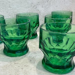 May include: A set of six emerald green drinking glasses. The glasses have a faceted design and a thick base. The glasses are made of a transparent material, allowing light to pass through. The glasses are arranged on a light-colored surface.