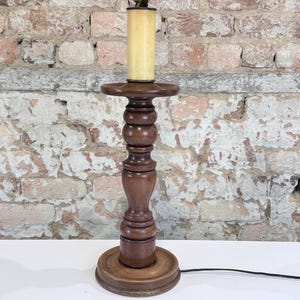 May include: A dark brown wooden table lamp base with a turned spindle design, featuring a circular base and a top platform. The lamp includes a cylindrical cream-colored socket cover and a metal light fixture component at the very top.