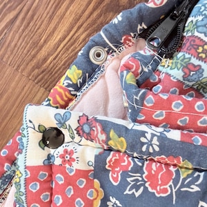 May include: A quilted fabric garment featuring a multi-colored floral and geometric patchwork pattern in shades of navy blue, red, yellow, and white. The piece includes a black YKK zipper, a black snap button, and a silver-toned metal snap fastener.