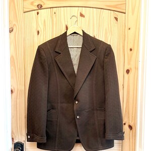 May include: A dark brown blazer with a textured fabric finish, featuring a notched lapel, two front buttons, and decorative button cuffs. The interior lining displays a white and black geometric pattern, and the garment is hung on a wooden hanger.