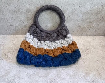 May include: A hand-knitted handbag with a circular handle. The bag features horizontal stripes of gray, white, tan, and blue. The knitted texture is visible throughout the bag's design, creating a cozy and textured appearance.