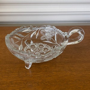 May include: A clear, decorative glass dish with a handle and three small feet. The dish features floral and geometric etched designs. The rim has a scalloped edge. The handle is a loop shape.