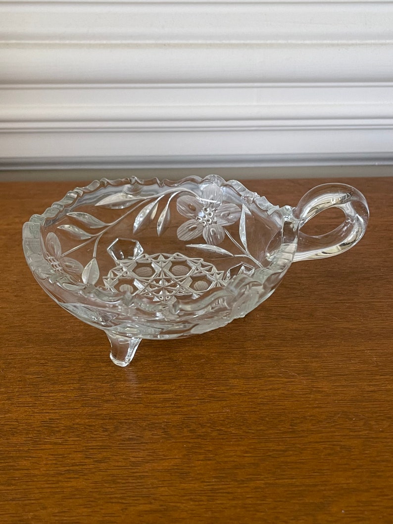 May include: A clear, decorative glass dish with a handle and three small feet. The dish features floral and geometric etched designs. The rim has a scalloped edge. The handle is a loop shape.