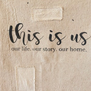 May include: A beige canvas wall hanging with the text "this is us our life. our story. our home." in black lettering. Two rectangular patches of fabric are sewn onto the canvas.