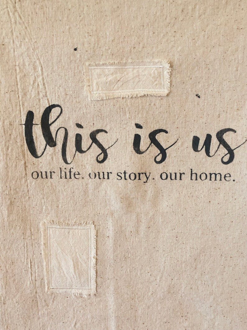 May include: A beige canvas wall hanging with the text "this is us our life. our story. our home." in black lettering. Two rectangular patches of fabric are sewn onto the canvas.