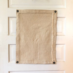 May include: A plain, off-white canvas banner with grommets in each corner. The banner is hanging on a white door.