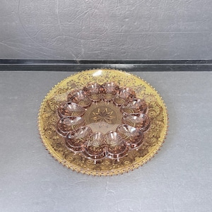 May include: A round amber-colored glass serving plate with an intricate embossed floral border and a serrated edge, featuring a central circular depression designed to hold twelve individual egg-shaped glass cups in a radial arrangement.