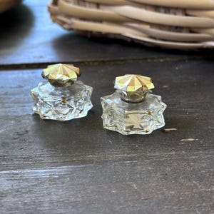 May include: Two small, clear glass perfume bottles with faceted sides and gold-colored geometric stoppers. The bottles sit on a dark wooden surface, with a woven basket in the background.