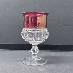 May include: A clear glass goblet with a ruby red rim and a decorative, patterned body. The goblet has a round base and a detailed stem. The rim has a metallic sheen, and the body features a repeating circular design.