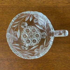 May include: Clear glass serving dish with a handle, featuring floral and geometric etched designs. The dish has a round shape with a handle on one side. The floral pattern includes flowers and leaves around the rim and center.