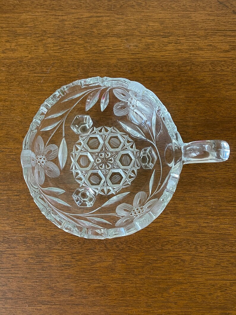 May include: Clear glass serving dish with a handle, featuring floral and geometric etched designs. The dish has a round shape with a handle on one side. The floral pattern includes flowers and leaves around the rim and center.