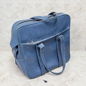May include: A blue satchel bag with a top handle and two shorter handles. The bag has a zipper closure and a small pocket on the side. The bag is made of a smooth material and has white stitching.