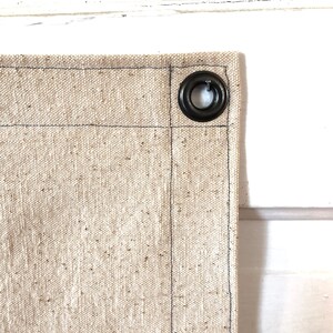 May include: Close-up of a beige canvas fabric with a black metal grommet. The fabric has a subtle texture and is sewn with black thread.