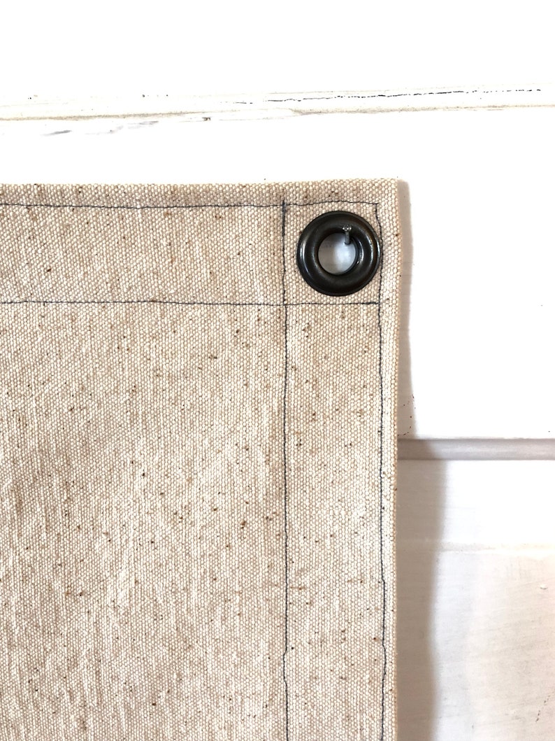 May include: Close-up of a beige canvas fabric with a black metal grommet. The fabric has a subtle texture and is sewn with black thread.