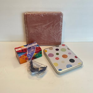 May include: An assortment of items including a wrapped, dusty rose-colored photo album, a colorful box, a polka-dot tin, and a small, dark blue item in a plastic bag. The items are arranged on a white surface.