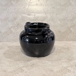 May include: A glossy black ceramic crock with a wide, rounded body and a short, thick neck. The vessel features two small, sturdy handles positioned on opposite sides of the rim. The interior is dark, and the exterior surface has a reflective finish.