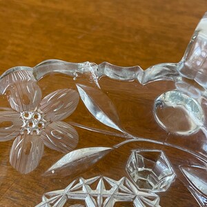May include: Close-up of a clear glass dish with floral and geometric etched designs. The dish features a flower, leaves, and a geometric pattern. The rim of the dish has a scalloped edge. The glass has a slight crack.