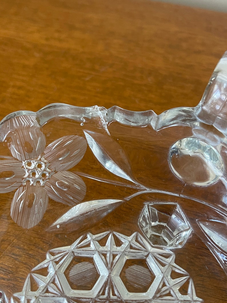 May include: Close-up of a clear glass dish with floral and geometric etched designs. The dish features a flower, leaves, and a geometric pattern. The rim of the dish has a scalloped edge. The glass has a slight crack.