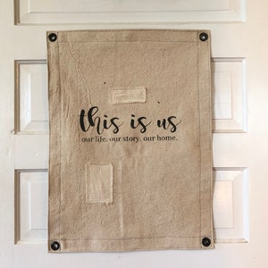 May include: A beige canvas wall hanging with the text "this is us our life, our story, our home." The canvas has two rectangular patches of fabric sewn on it.