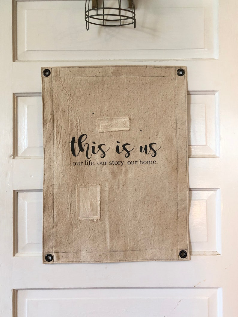 May include: A beige canvas wall hanging with the text "this is us our life, our story, our home." The canvas has two rectangular patches of fabric sewn on it.