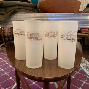 May include: Four frosted glass tumblers with brown illustrations of trucks and construction vehicles. The glasses are cylindrical and sit on a wooden stool with a dark brown finish. The background includes a purple patterned rug.