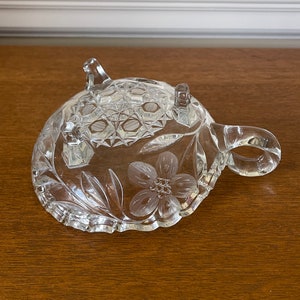 May include: A clear, decorative glass dish with a floral design and three small feet. The dish has a handle and a textured pattern on the top surface. The floral design is etched into the glass, adding a delicate touch.