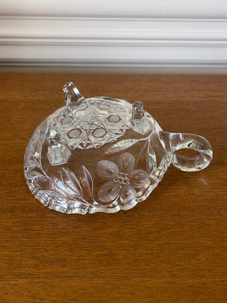 May include: A clear, decorative glass dish with a floral design and three small feet. The dish has a handle and a textured pattern on the top surface. The floral design is etched into the glass, adding a delicate touch.