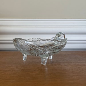 May include: A clear, decorative glass dish with a scalloped rim and three small feet. The dish has a floral pattern etched into the glass and a small handle. The dish is approximately 5 inches long and 3 inches wide.