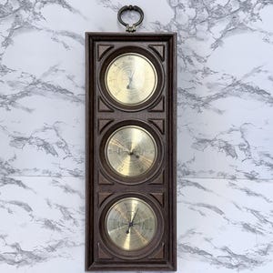May include: A vintage weather station with three brass-faced gauges set in a dark brown wooden frame. The gauges are arranged vertically, with a metal loop at the top for hanging. The background is a white marble pattern.