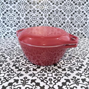 May include: A glossy red enameled cast iron Dutch oven with a matching lid. The pot features two integrated side handles and a rounded, deep basin design. The exterior surface is smooth and solid red, showcasing a sturdy construction for cooking.