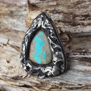 May include: A silver ring with a turquoise stone set in a rough, textured metal setting. The ring is shaped like a triangle and has a rustic, handmade look.