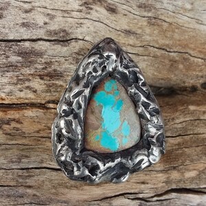 May include: A triangular silver ring with a turquoise stone set in the center. The ring has a textured, rough surface.