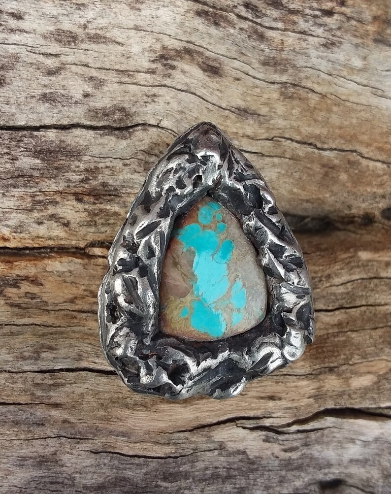 May include: A triangular silver ring with a turquoise stone set in the center. The ring has a textured, rough surface.