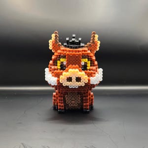 May include: A brown and white bead-based figurine of a warthog with a black hat. The figurine has yellow eyes and a white snout.