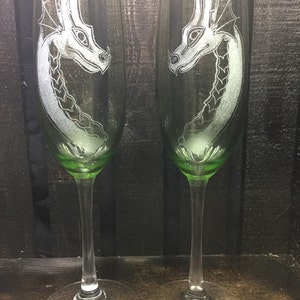 May include: Two clear glass champagne flutes with green stems and bases. Each flute has an etched design of a white dragon's head.