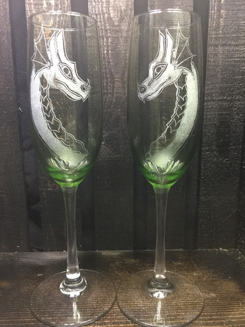 May include: Two clear glass champagne flutes with green stems and bases. Each flute has an etched design of a white dragon's head.