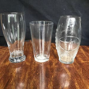 May include: Three clear glass drinking glasses of different shapes and sizes. The glasses are on a wooden surface.