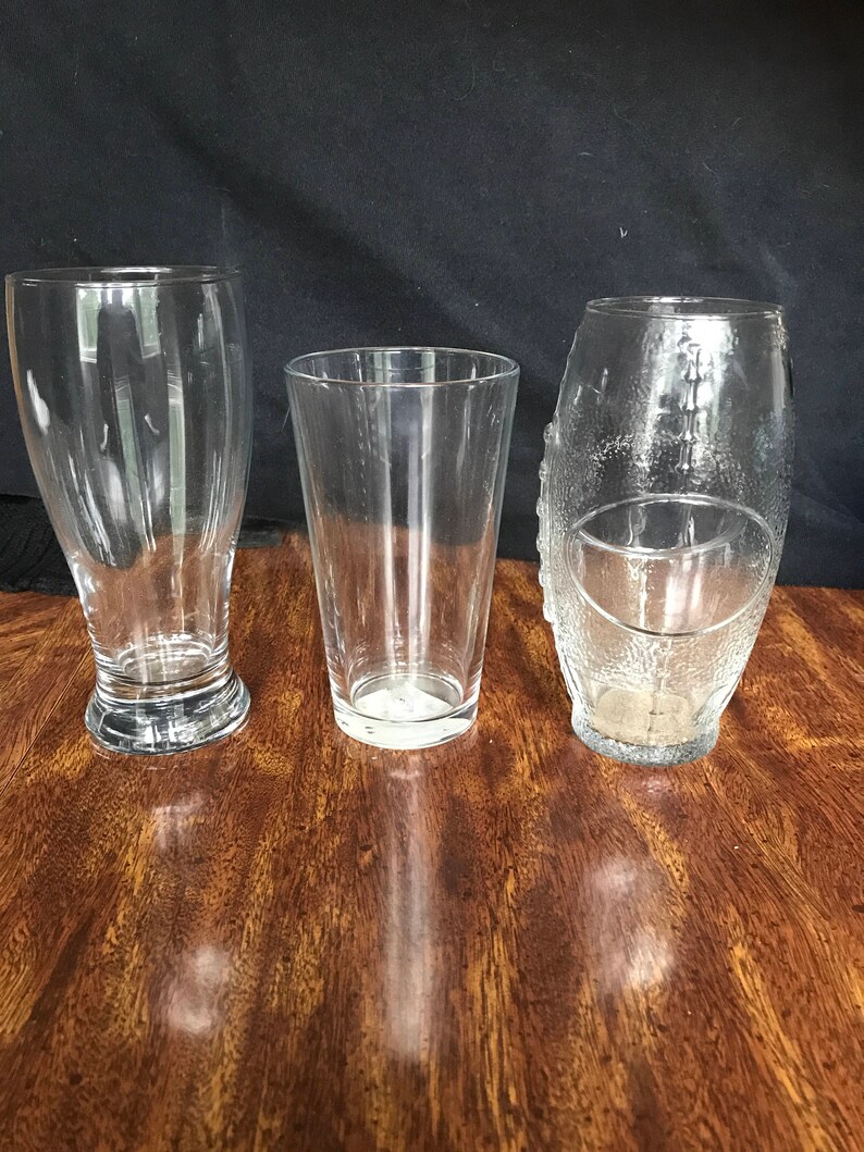 May include: Three clear glass drinking glasses of different shapes and sizes. The glasses are on a wooden surface.