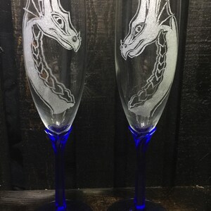 May include: Two clear glass champagne flutes with blue stems and bases. Each flute is etched with a white dragon design.
