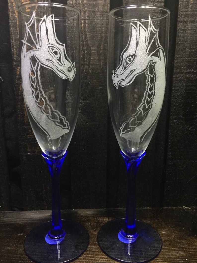 May include: Two clear glass champagne flutes with blue stems and bases. Each flute is etched with a white dragon design.